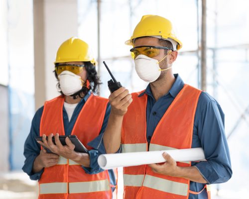 Two construction workers wearing safety vests, hard hats, and face masks, holding blueprints and a walkie-talkie at a construction site.