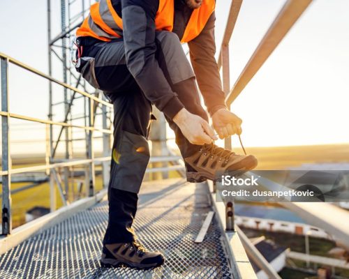 An industry worker tying shoelace on work shoes while standing on metal construction.
